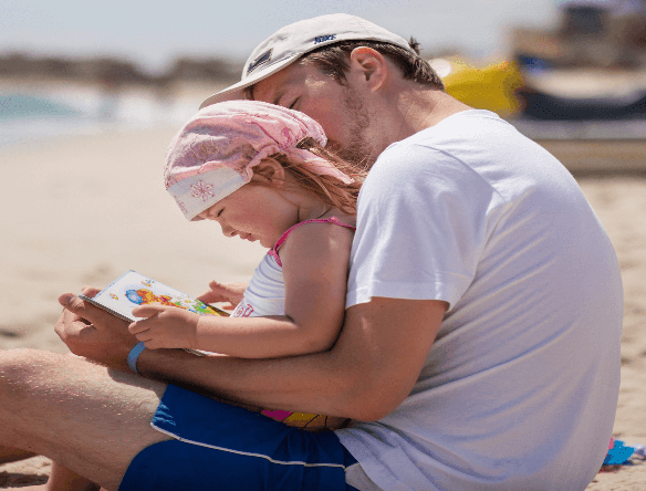 A man reading a book to a child on the beach